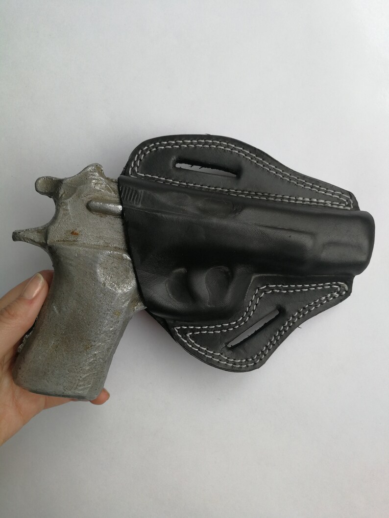 Puede incluir: Una funda de cuero negra con costuras blancas para una pistola. La funda est&aacute; dise&ntilde;ada para ajustarse perfectamente al arma y tiene una solapa que cubre el gatillo.