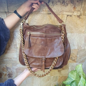 Peut inclure: Un sac &agrave; bandouli&egrave;re en cuir marron avec une cha&icirc;ne dor&eacute;e. Le sac comporte plusieurs compartiments zipp&eacute;s et une poche avant &agrave; rabat. Le sac est tenu par les mains d'une personne.