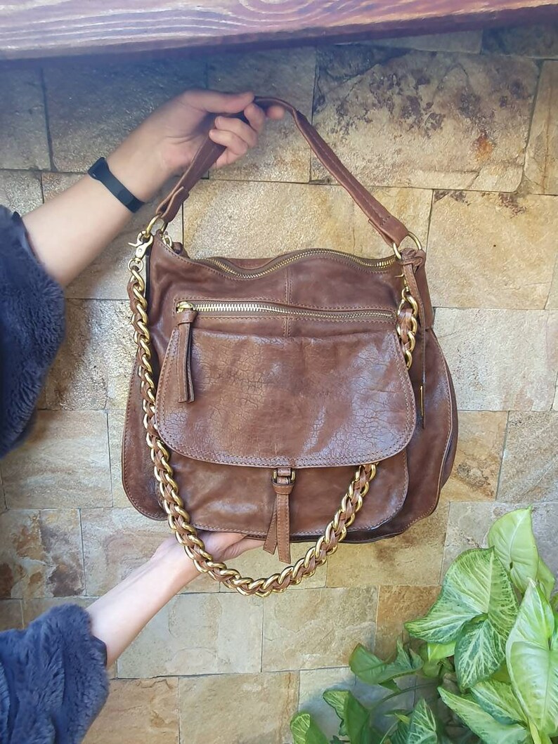 Peut inclure: Un sac &agrave; bandouli&egrave;re en cuir marron avec une cha&icirc;ne dor&eacute;e. Le sac comporte plusieurs compartiments zipp&eacute;s et une poche avant &agrave; rabat. Le sac est tenu par les mains d'une personne.