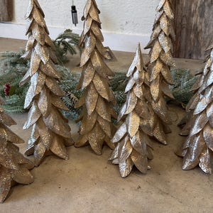 May include: A collection of seven decorative tabletop trees featuring a metallic gold finish and a layered leaf-like texture. Each tree has a pointed conical shape with overlapping petal structures that create a dense, textured appearance.