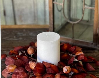 May include: A white pillar candle sits in the center of a square wooden base, surrounded by a wreath of deep red eucalyptus leaves and small brown seed pods.