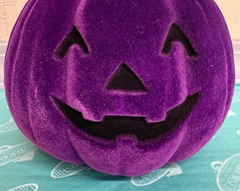 May include: A plush, purple Halloween pumpkin with a carved jack-o'-lantern face. The pumpkin has a soft, velvety texture and a brown stem. The smiling face features triangular eyes and a wide, toothy grin. A festive decoration for the fall season.