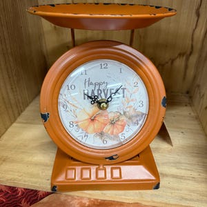 May include: Orange decorative clock in the shape of a vintage scale. The clock face reads "Happy Harvest" with pumpkin illustrations. The clock has a distressed finish, and the scale tray is at the top.