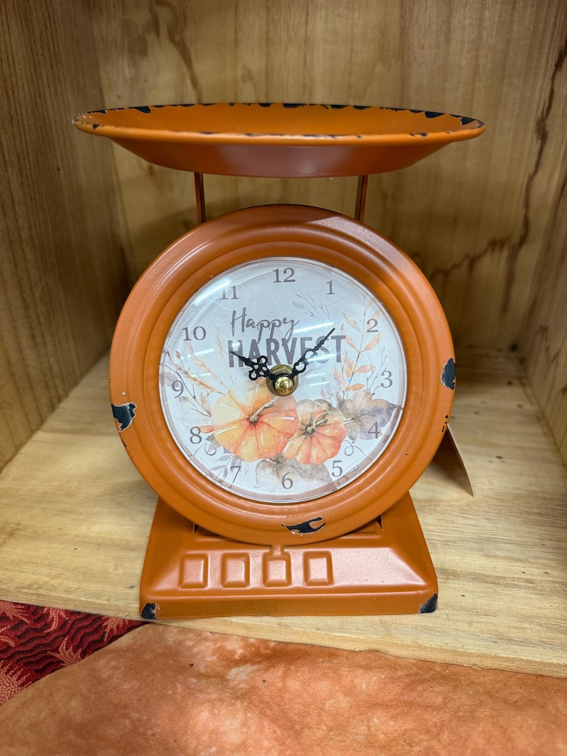 May include: Orange decorative clock in the shape of a vintage scale. The clock face reads "Happy Harvest" with pumpkin illustrations. The clock has a distressed finish, and the scale tray is at the top.