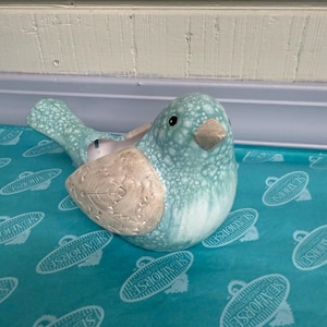 May include: A ceramic bird-shaped planter in shades of blue and white. The bird has a textured finish and a beige wing detail. The planter is sitting on a teal surface with a repeating pattern of white seahorse logos.