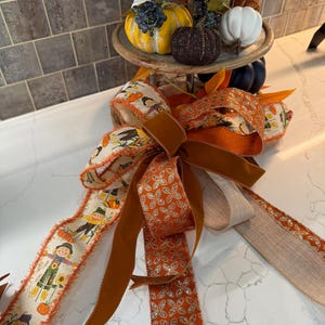 May include: A fall-themed ribbon with a brown velvet ribbon, a gold glitter ribbon with a floral pattern, and a white ribbon with a brown trim and a pattern of scarecrows and pumpkins.