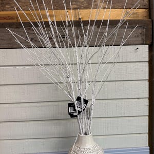 May include: A white decorative birch branch arrangement in a textured, off-white vase. The branches are thin and numerous, creating a full, airy display. The vase has a dotted pattern. A small white device with a cord is visible.