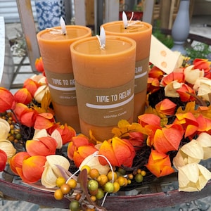 May include: Three orange pillar candles with the text "TIME TO RELAX" on a brown label. The candles are surrounded by a wreath of orange, yellow, and green fall foliage, berries, and leaves. The candles are on a round, dark metal table.