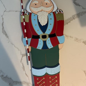 May include: Wooden Christmas nutcracker soldier decoration. The figure is painted in red, blue, green, and gold. It holds a candy cane-striped staff with a red and white swirl lollipop. The nutcracker has a white beard and mustache.