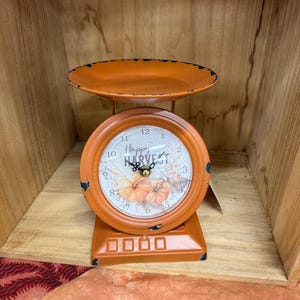 May include: Orange decorative scale with a round clock face. The clock face reads "Happy Harvest" and features pumpkin illustrations. The scale has a small tray and a rectangular base. The scale is made of metal.