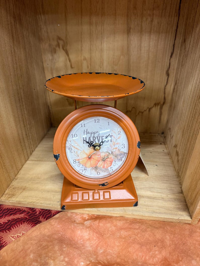 May include: Orange decorative scale with a round clock face. The clock face reads "Happy Harvest" and features pumpkin illustrations. The scale has a small tray and a rectangular base. The scale is made of metal.