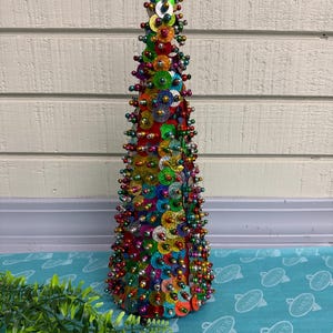 May include: A colorful, cone-shaped decorative tree covered in sequins and small beads. The tree features a variety of colors, including red, green, blue, and gold. The tree is approximately 12 inches tall and is a festive holiday decoration.