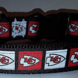 May include: Black dog collar featuring a repeating pattern of red and white squares with the Kansas City Chiefs logo. The collar has a black plastic buckle and a metal D-ring for leash attachment. The collar is made of a durable, woven material.