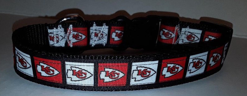 May include: Black dog collar featuring a repeating pattern of red and white squares with the Kansas City Chiefs logo. The collar has a black plastic buckle and a metal D-ring for leash attachment. The collar is made of a durable, woven material.