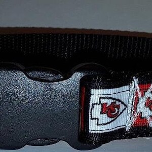 May include: Black dog collar featuring a repeating pattern of the Kansas City Chiefs logo in red, white, and gold. The collar has a black plastic buckle and a metal D-ring for leash attachment. The collar is approximately 1 inch wide.