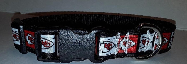 May include: Black dog collar featuring a repeating pattern of the Kansas City Chiefs logo in red, white, and gold. The collar has a black plastic buckle and a metal D-ring for leash attachment. The collar is approximately 1 inch wide.
