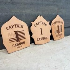 May include: Three light tan leather firefighter helmet shields with dark brown laser-engraved text and graphics. The left shield displays CAPTAIN, CANNON, HCFR, and a flag. The center shield shows CAPTAIN, 1, and CANNON. The right shield reads FIREFIGHTER, CANNON, and HCFR.
