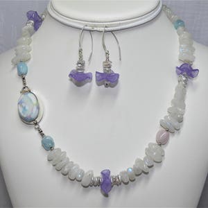 May include: A white and blue gemstone necklace with lavender flower accents. The necklace is made with moonstone and aquamarine beads. The earrings are also made with lavender flower accents and moonstone.
