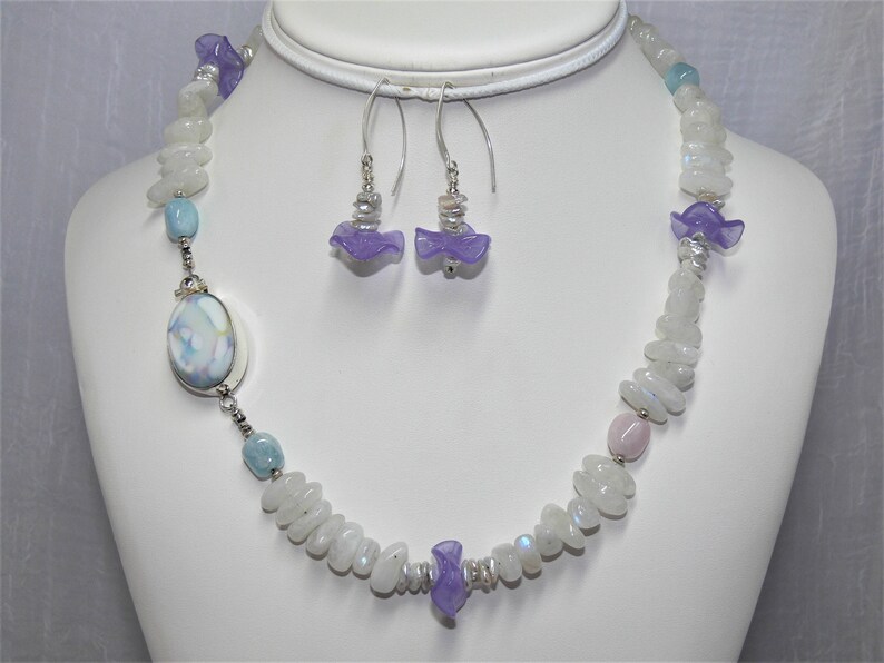May include: A white and blue gemstone necklace with lavender flower accents. The necklace is made with moonstone and aquamarine beads. The earrings are also made with lavender flower accents and moonstone.
