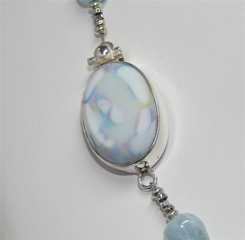 May include: A silver necklace with a large oval pendant featuring a blue and white marbled glass design. The pendant is attached to a silver chain with smaller beads.