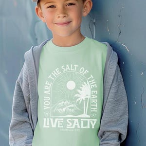 Op de afbeelding: Een lichtgroen T-shirt met een wit grafisch ontwerp met een zon, palmbomen en golven. De tekst "You are the salt of the earth" staat in een cirkel rond de afbeelding. Onder de afbeelding staat de tekst "Live Salty" met de woorden "Matthew 5:13" eronder.