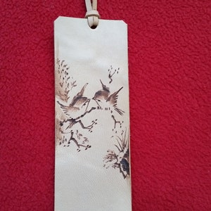 Puede incluir: Un marcap&aacute;ginas de cuero marr&oacute;n claro con un dise&ntilde;o quemado de dos p&aacute;jaros posados en una rama.