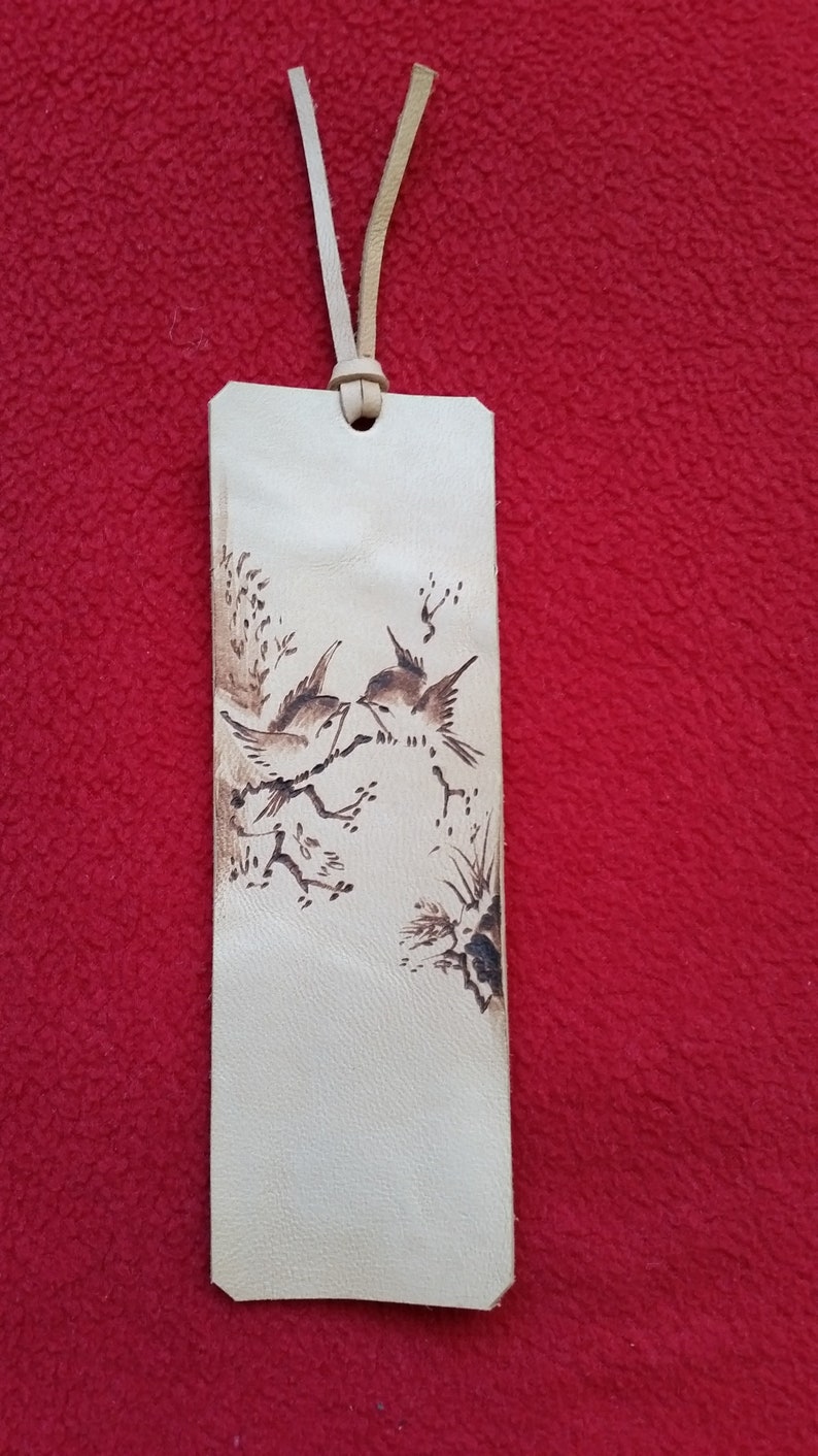 Puede incluir: Un marcap&aacute;ginas de cuero marr&oacute;n claro con un dise&ntilde;o quemado de dos p&aacute;jaros posados en una rama.