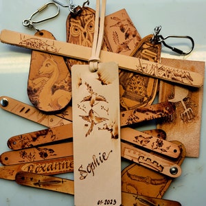 Puede incluir: Una colecci&oacute;n de marcap&aacute;ginas de cuero con varios dise&ntilde;os, incluyendo p&aacute;jaros, b&uacute;hos y patrones florales. Los marcap&aacute;ginas est&aacute;n todos quemados a mano y presentan una est&eacute;tica r&uacute;stica &uacute;nica. Un marcap&aacute;ginas est&aacute; personalizado con el nombre "Sophie" y la fecha "01-2023".