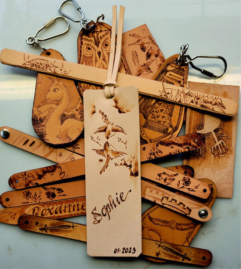 Puede incluir: Una colecci&oacute;n de marcap&aacute;ginas de cuero con varios dise&ntilde;os, incluyendo p&aacute;jaros, b&uacute;hos y patrones florales. Los marcap&aacute;ginas est&aacute;n todos quemados a mano y presentan una est&eacute;tica r&uacute;stica &uacute;nica. Un marcap&aacute;ginas est&aacute; personalizado con el nombre "Sophie" y la fecha "01-2023".