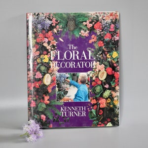 Op de afbeelding: Een paars boek getiteld "The Floral Decorator" van Kenneth Turner. De omslag toont een kleurrijke bloemstuk met een persoon in het midden.