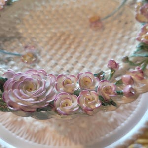 May include: A white ceramic plate with a delicate, openwork design. The rim is adorned with detailed, three-dimensional pink and white roses with green leaves. The plate has a scalloped edge.