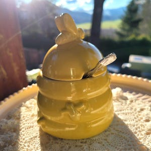 May include: A yellow ceramic honey pot shaped like a beehive, with a bee-shaped handle on the lid. A small silver spoon rests inside. The pot sits on a decorative, off-white lace doily.