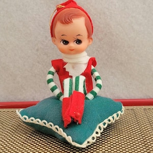 May include: A vintage Christmas elf doll with a red hat and outfit, green and white striped sleeves, and red boots. The doll sits on a teal pillow with a white scalloped edge. A gold string is attached to the hat.