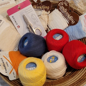 May include: A collection of colorful candlewicking yarn balls in red, blue, yellow, orange, and white, alongside packaged Singer folding scissors and various spools of white and light blue cotton thread, all arranged within a woven basket.