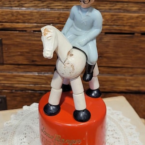 May include: Vintage Lone Ranger toy figurine. The toy features a white horse with a rider in a light blue outfit and hat. The figurine is mounted on a red base with text. The toy is made of plastic and has a classic, nostalgic appeal.