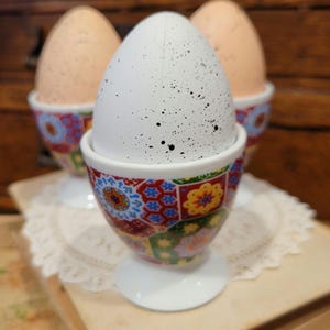 May include: Three eggs in decorative egg cups. One white egg with black speckles sits in a colorful cup with floral and geometric patterns. Two brown eggs are in similar cups. The cups are on a white doily and books.