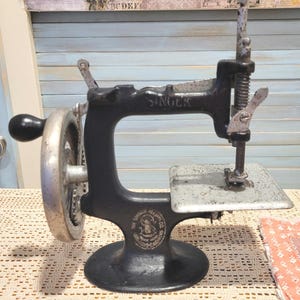 May include: A vintage Singer sewing machine, painted black, with a silver hand wheel and needle assembly. The machine has the Singer logo and the words "The Singer Manufacturing Co." on the base. The machine sits on a cream-colored crocheted surface.