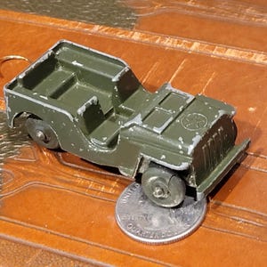 May include: A vintage olive green toy jeep with silver accents. The toy jeep has a detailed design, including a front grill, headlights, and a star emblem on the hood. The jeep is approximately 3 inches long.