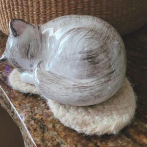 May include: A ceramic figurine of a sleeping cat with a white and grey brushstroke pattern, featuring pink accents on the ears and nose. The cat is curled into a ball and rests on a small, oval-shaped, cream-colored crocheted fabric mat.