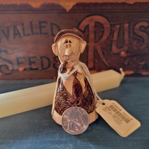 May include: A handcrafted ceramic figurine of a man wearing a cap, scarf, and vest. The figurine is brown and tan with intricate details. A quarter is placed in front of the figurine for scale. A tag is attached to the figurine.