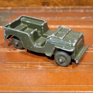 May include: An olive green toy jeep with a weathered appearance. The miniature vehicle has four wheels, a front bumper, and a detailed interior. The paint shows signs of wear, suggesting it is a vintage item.