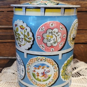 May include: Vintage blue tin canister with a lid. The canister is decorated with colorful plates featuring floral and bird designs. The lid and top rim are also blue. A decorative storage container for the kitchen.