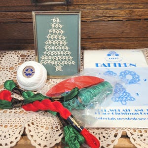 May include: A framed, off-white lace Christmas tree on a teal background. A ball of white DMC thread, red and green embroidery floss, and a pattern for lace Christmas ornaments are also visible. The items are arranged on a lace doily.