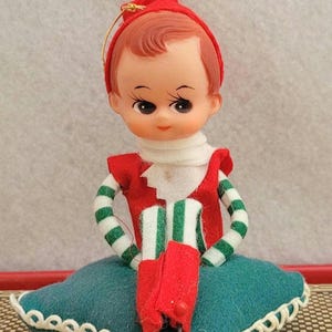 May include: A small elf figurine with a molded plastic face, wearing a red hat, a red vest, and white and green striped sleeves. The elf sits on a square teal felt pillow with white scalloped trim, featuring a gold hanging loop on the hat.