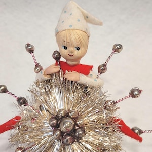 May include: A vintage elf figurine with blonde hair and a white hat featuring blue polka dots. The elf wears a red collar and is positioned behind a large, spherical silver tinsel ornament decorated with metallic beads on thin wire stems.