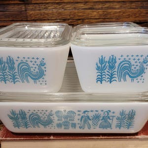 May include: A set of vintage white Pyrex dishes with clear glass lids. The dishes feature a blue rooster and floral design. The set includes two square containers and a rectangular baking dish, stacked on top of each other.