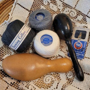 May include: A collection of sewing supplies including a black ball of darning cotton, a grey ball of darning cotton, a white ball of darning cotton, a black wooden darning egg, a light brown wooden darning mushroom, and a pack of Boye needles.