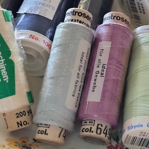 May include: Close-up of several spools of thread in various colors, including blue, lavender, and mint green. The spools have labels with text in multiple languages, including "Polyester spun" and "Ideal for all fabrics."