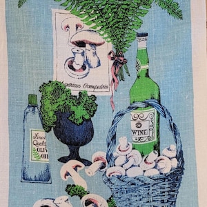 May include: A vintage-style kitchen towel with a light blue background featuring a still life illustration. The image includes a basket of mushrooms, a green wine bottle, a bottle of olive oil, and a fern. The towel has a rustic, hand-drawn aesthetic.