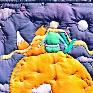 Puede incluir: Un tapiz de pared de tela acolchada azul con un círculo amarillo en el centro. El círculo tiene una oveja blanca y el texto "B 612" está debajo. El fondo es azul oscuro con estrellas y nubes.